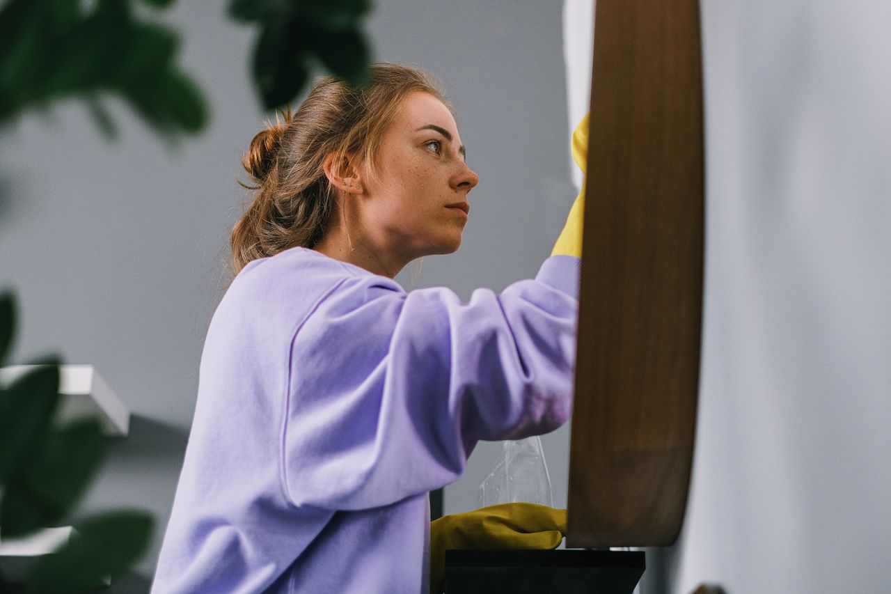 Vue latérale d’une jeune femme concentrée portant des gants en latex, debout et nettoyant un miroir dans une pièce lumineuse en plein jour